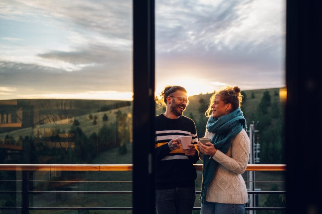 Affectionate young couple drinking coffee or tea while standing on their balcony at home in the morning with a beautiful nature view. Watching the sunrise with a romantic partner. Copy space.