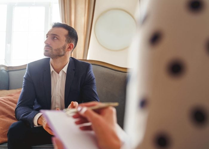 Frustrated man with a short haircut looking away Joyless elegant young male patient sitting on a couch in front of a female psychologist